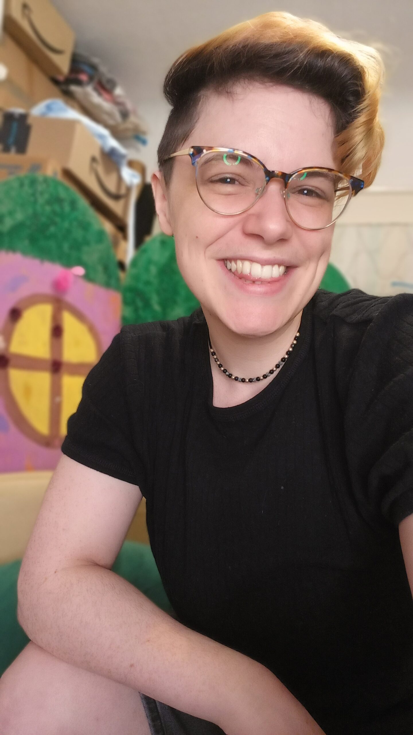 A slim white human is leaning slightly forward to the camera, with a big grin on their face. They have short blonde hair with dark brown roots which is shaved at the sides. They are wearing a black t-shirt, a small black beaded necklace, and horn-rimmed glasses.