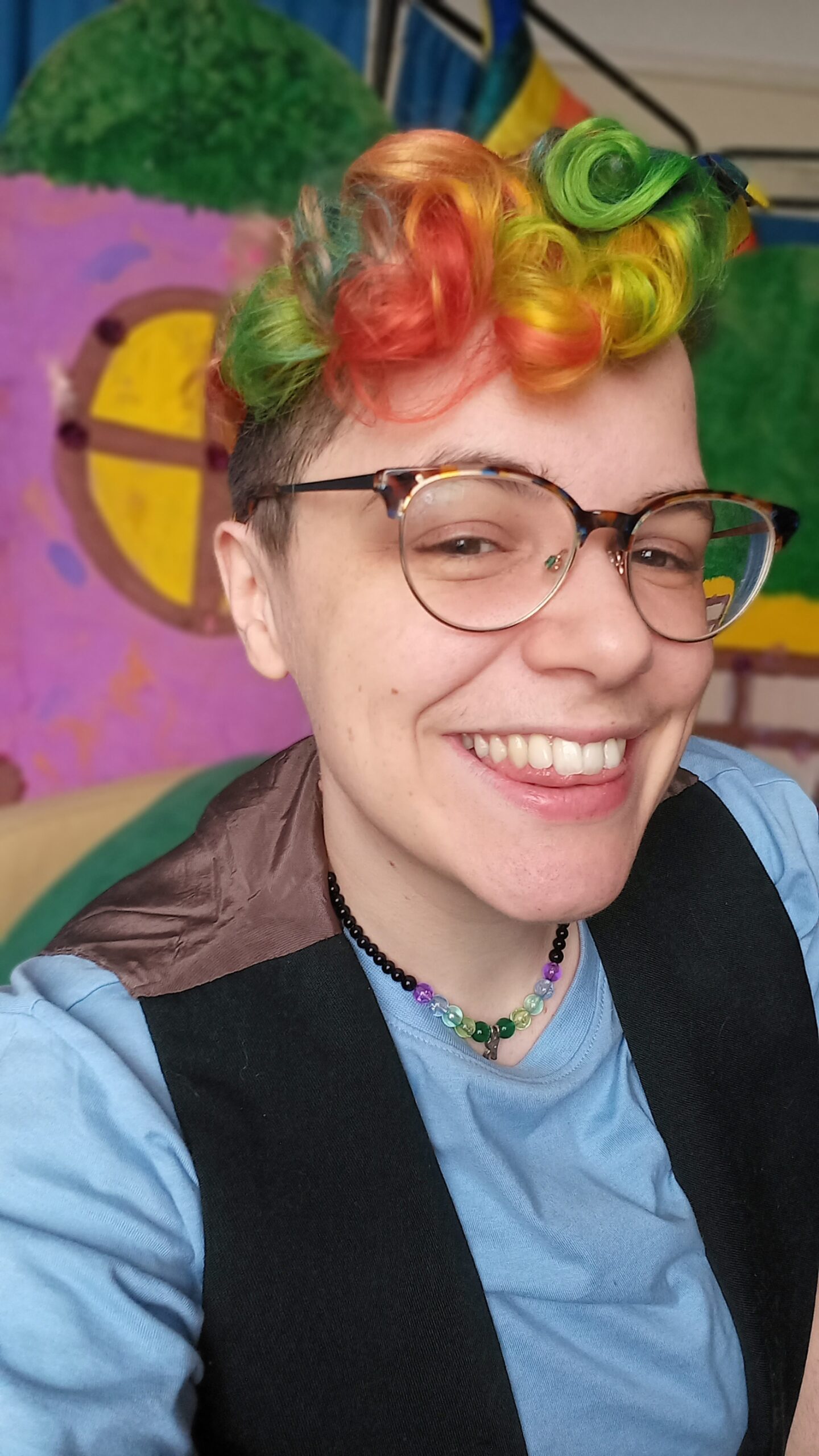 A slim white human, leaning forwards to the camera, smiling joyfully showing their teeth. They are wearing round horn rimmed glasses and have vivid rainbow coloured curly hair on top of their head and shaved sides. They are wearing a sky blue t-shirt with a black waistcoat, and a necklace with black, purple, blue, and green beads. Behind them is a painted backdrop which is bright pink with a circular painted window in yellow and brown.