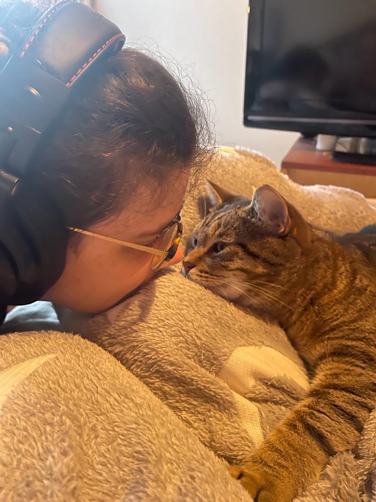A tabby cat laying on a grey fluffy blanket, nose to nose with Ren who is wearing over the ear headphones and glasses.