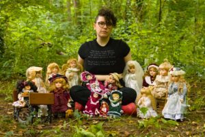 Vox is sitting cross legged in a clearing in a forest, staring at the camera with a blank expression on their face. Their short dark curls are falling over their forehead on one side, and on top of their round glasses. They are surrounded by a range of vintage porcelain dolls. Some are on their lap, and others are positioned closely next to and in front of them.