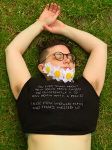 Vox is laying on grass with their eyes closed, and their arms above their head, with one of their hands gently resting in the other. They have dark brown underarm hair, and are wearing a beard made from embroidered marguerite flowers. The text on their cropped t-shirt can be clearly read. 