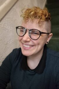 A white nonbinary person with short curly bleached hair on top and shaved sides. They are beaming, showing their cheek dimples and laughter lines around their eyes. They are wearing a black stud earring, a dark green jumper over a black collared t-shirt, and large round glasses with a brown frame and silver swirly decor on the lenses near the nose bridge. 