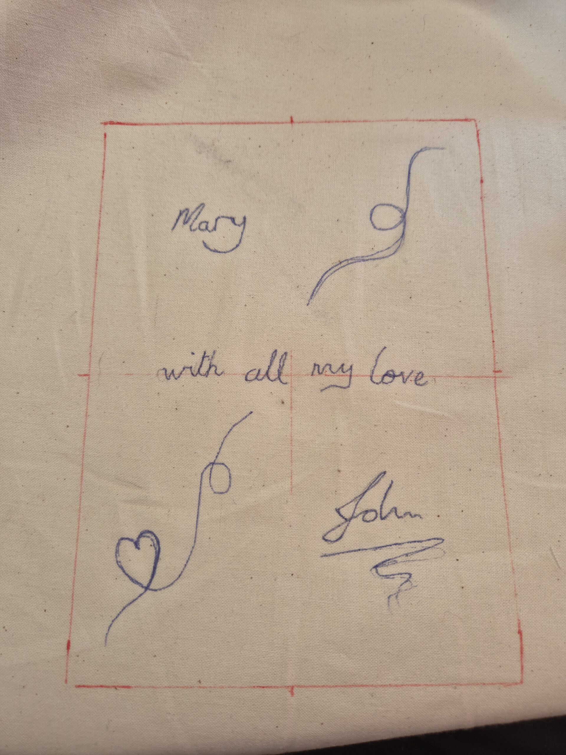 White fabric with a red rectangle drawn on it and the text "Mary with all my love John" and some decorative swirls drawn inside the rectangle.