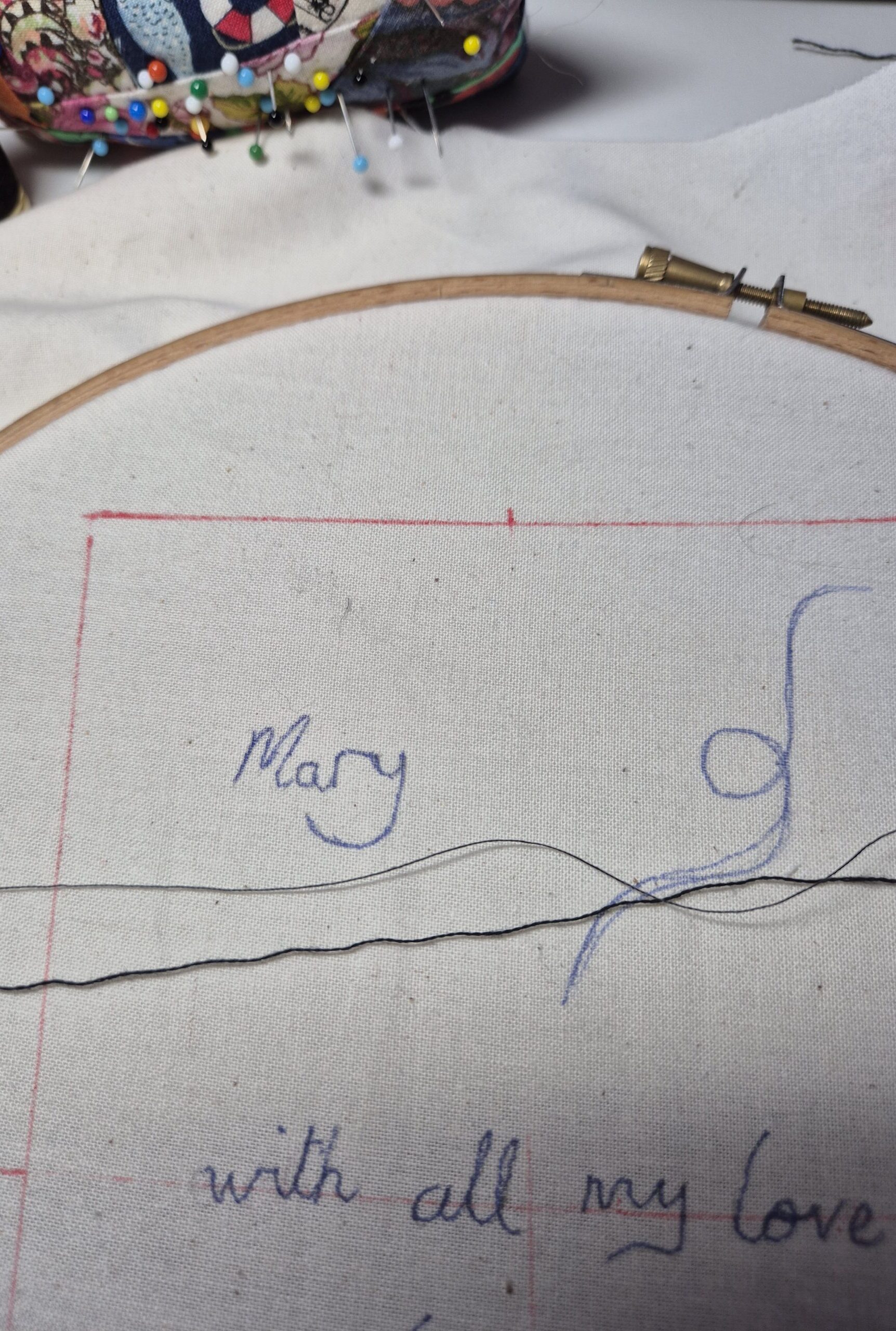 White fabric in an embroidery hoop. the words "Mary with all my love" are written on the fabric and there are two black threads laying across the fabric, one thin and one thicker