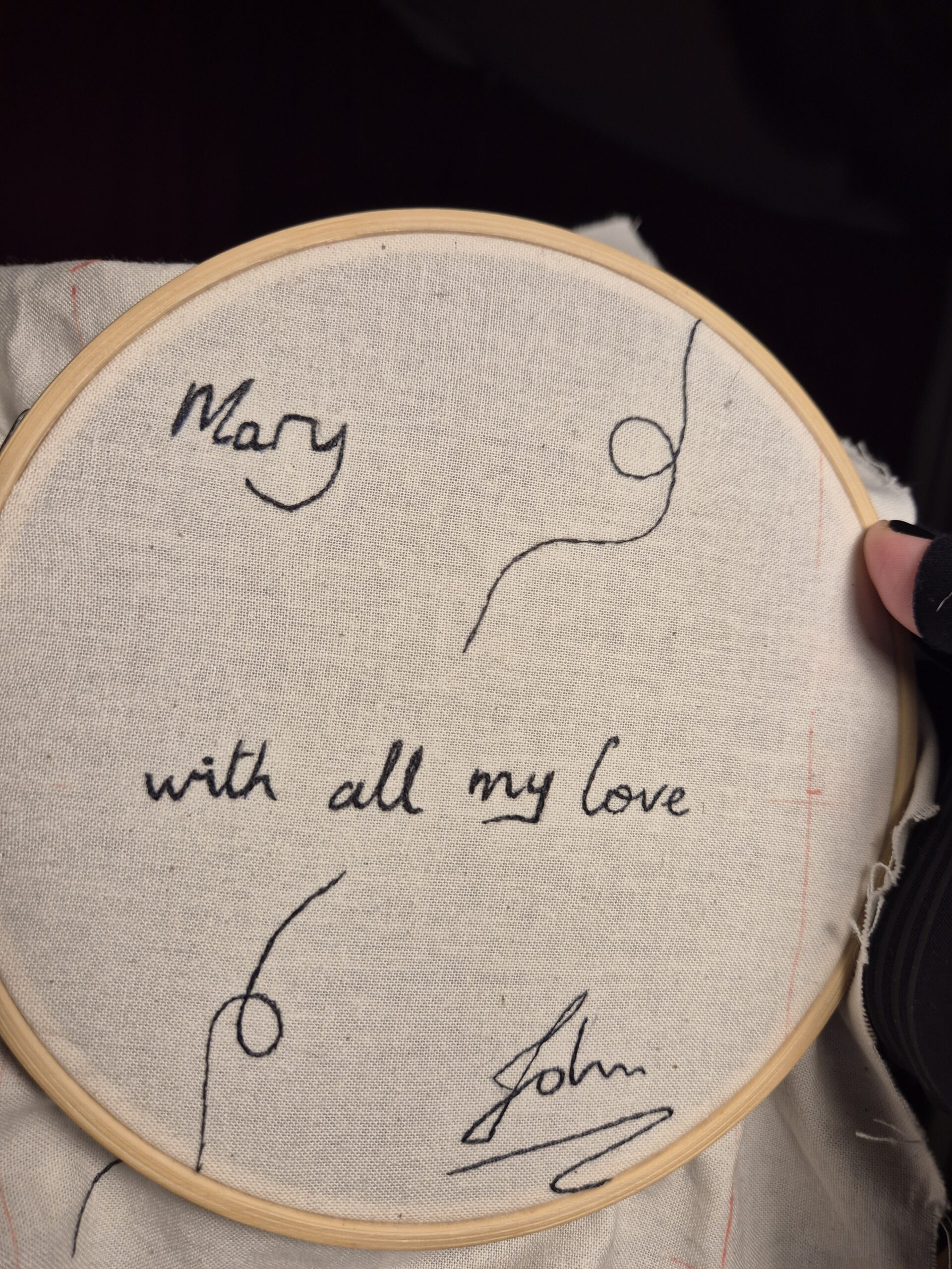 White fabric in an embroidery hoop. the words "Mary with all my love John" and some decorative swirls are embroidered on the fabric in black thread.