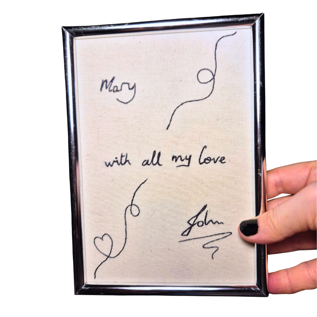 A silver frame with and embroidered piece of art in it with the words: "Mary with all my love John" with some decorative swirls around them.